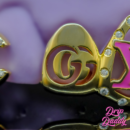 Drip Daddy Cut-Out Grillz in gold with gemstone accents, featuring a GG logo design. Displayed on a purple-black background with the Drip Daddy watermark.