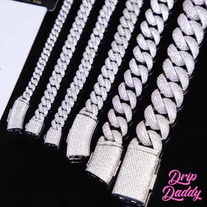 A person wearing Drip Daddy’s Frosted Miami Cuban Link Chain in 12mm, crafted in white gold plating with moissanite stones, layered in multiple lengths over a black shirt, with the Drip Daddy logo in pink at the bottom right.