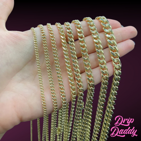 Close-up of a hand holding multiple sizes of Drip Daddy’s The Classic Miami Cuban Link Chains in yellow gold plated finish, ranging from 3mm to 18mm thickness, displayed against a burgundy background, with the Drip Daddy logo in pink at the bottom right corner.