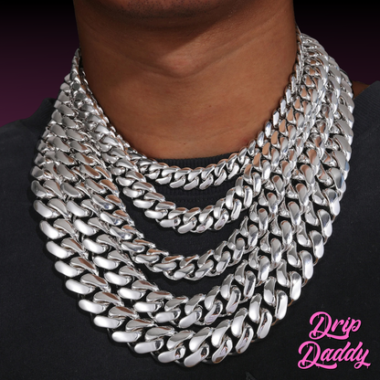A person wearing Drip Daddy’s The Don – 20mm to 30mm Miami Cuban Link Chain in a white gold plated finish, styled in a layered stack around the neck on a black shirt, with the Drip Daddy logo in pink visible in the bottom right corner.