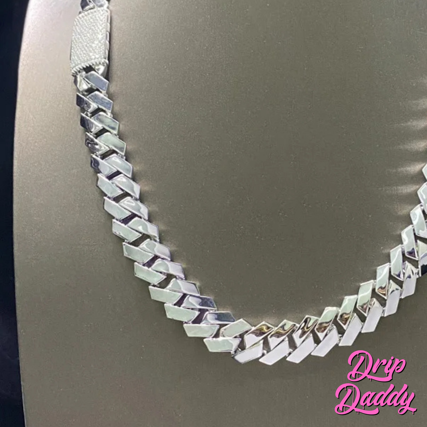 Close-up of Drip Daddy’s The Hustler Cuban Link Chain in white gold plated finish displayed against a grey background, highlighting the polished links and iced clasp, with the pink Drip Daddy logo visible in the bottom right corner.