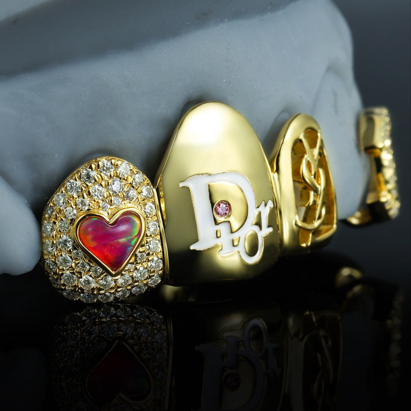 Custom luxury gold grillz set featuring a red opal heart surrounded by diamonds, Dior logo tooth with pink diamond accent, and YSL design. Handcrafted by Drip Daddy for a bold and high-end statement look