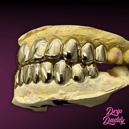 A plaster dental mold showcases Solid Plain Grillz in polished yellow gold, reflecting light with a mirror finish. The setup rests against a dark purple gradient backdrop with the Drip Daddy logo in pink cursive at the corner.