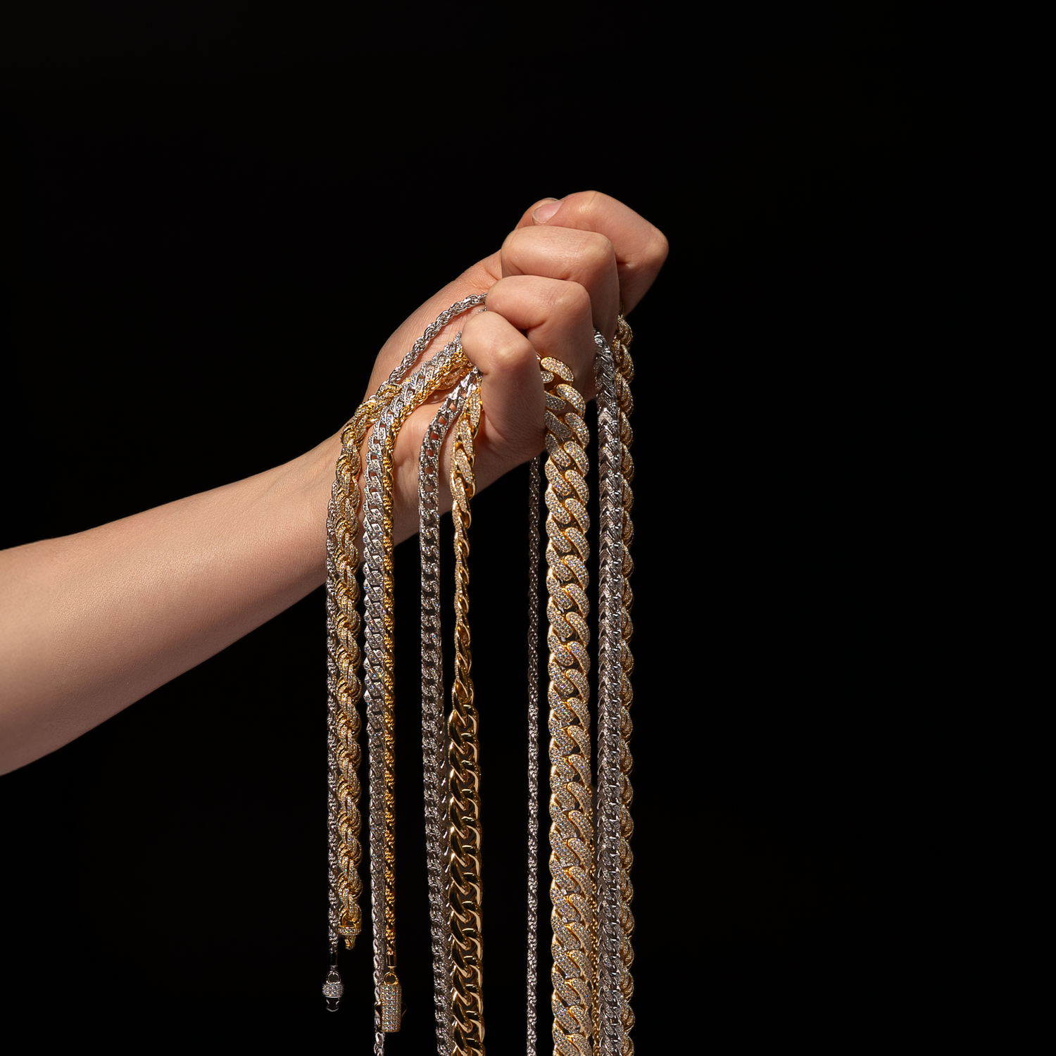 A hand holding multiple iced-out gold and silver chains, including rope, Cuban link, and tennis styles, showcasing luxury hip-hop jewelry by Drip Daddy against a black background.